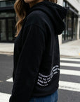 Person wearing a black hoodie with text on the back, standing on a street.