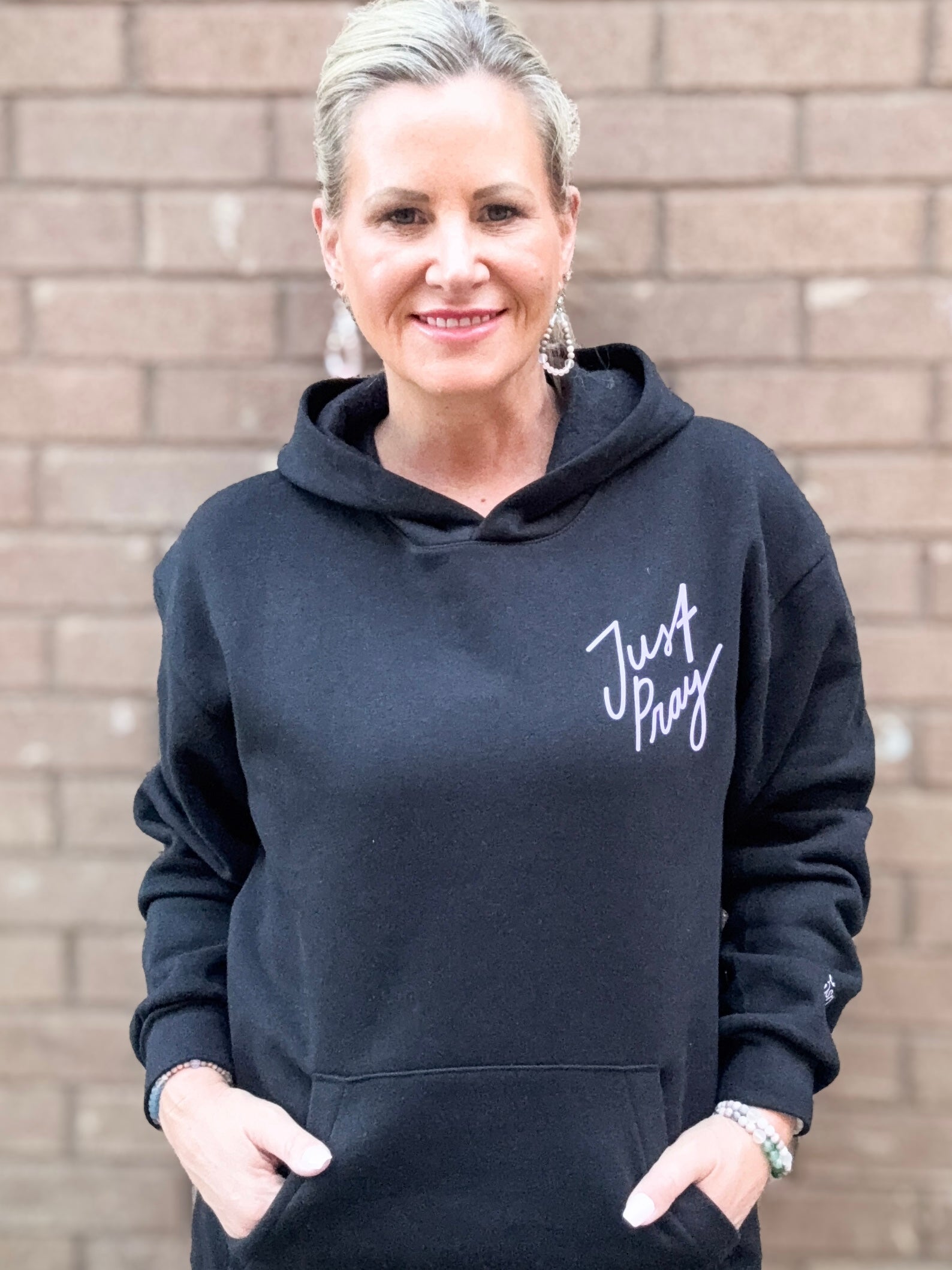 Woman wearing a black hoodie with 'Just Pray' text against a brick wall.