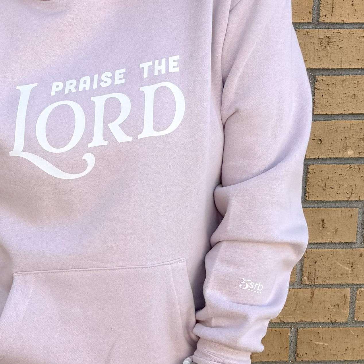Light pink hoodie with 'Praise the Lord' text against a brick wall.