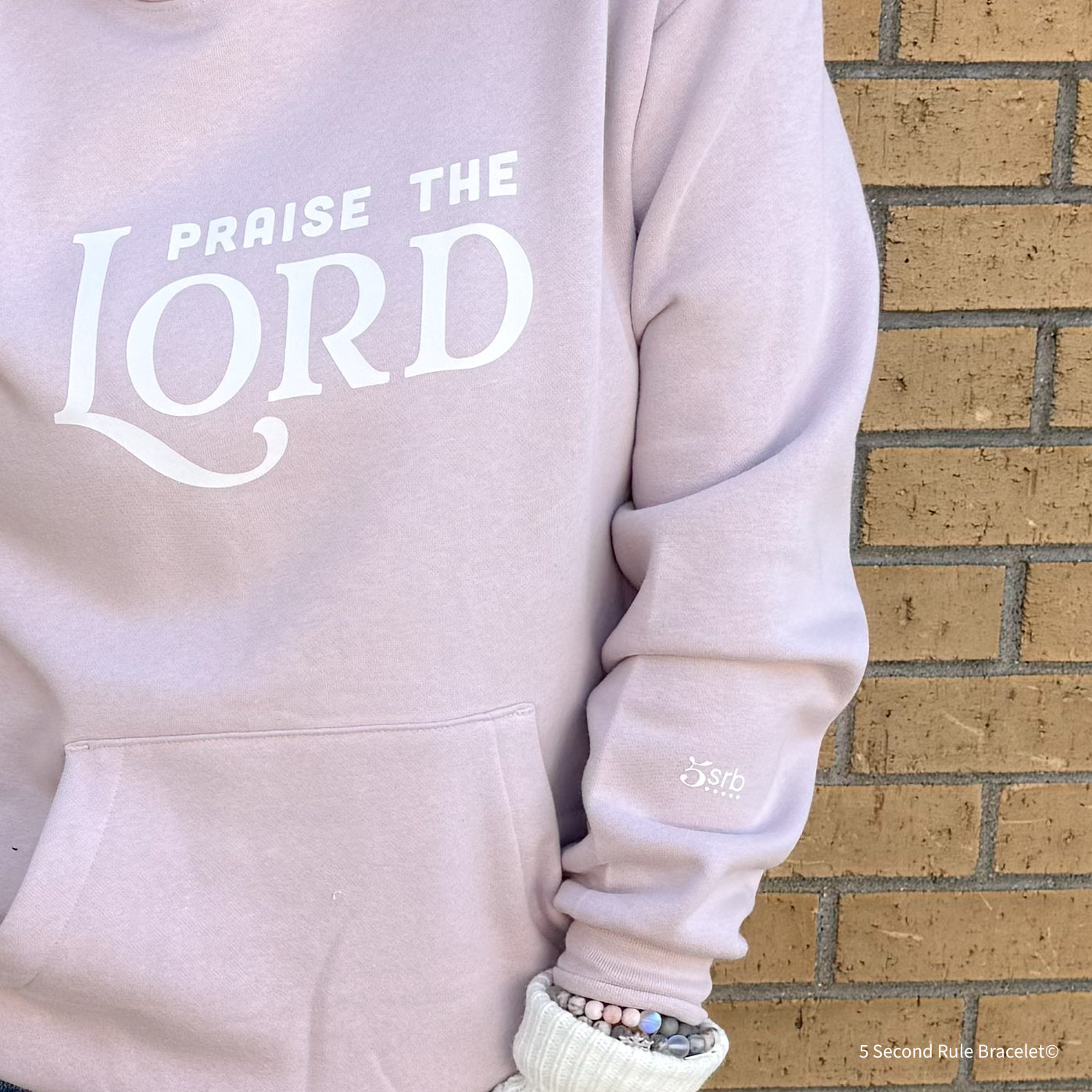 Light pink hoodie with 'Praise the Lord' text against a brick wall.