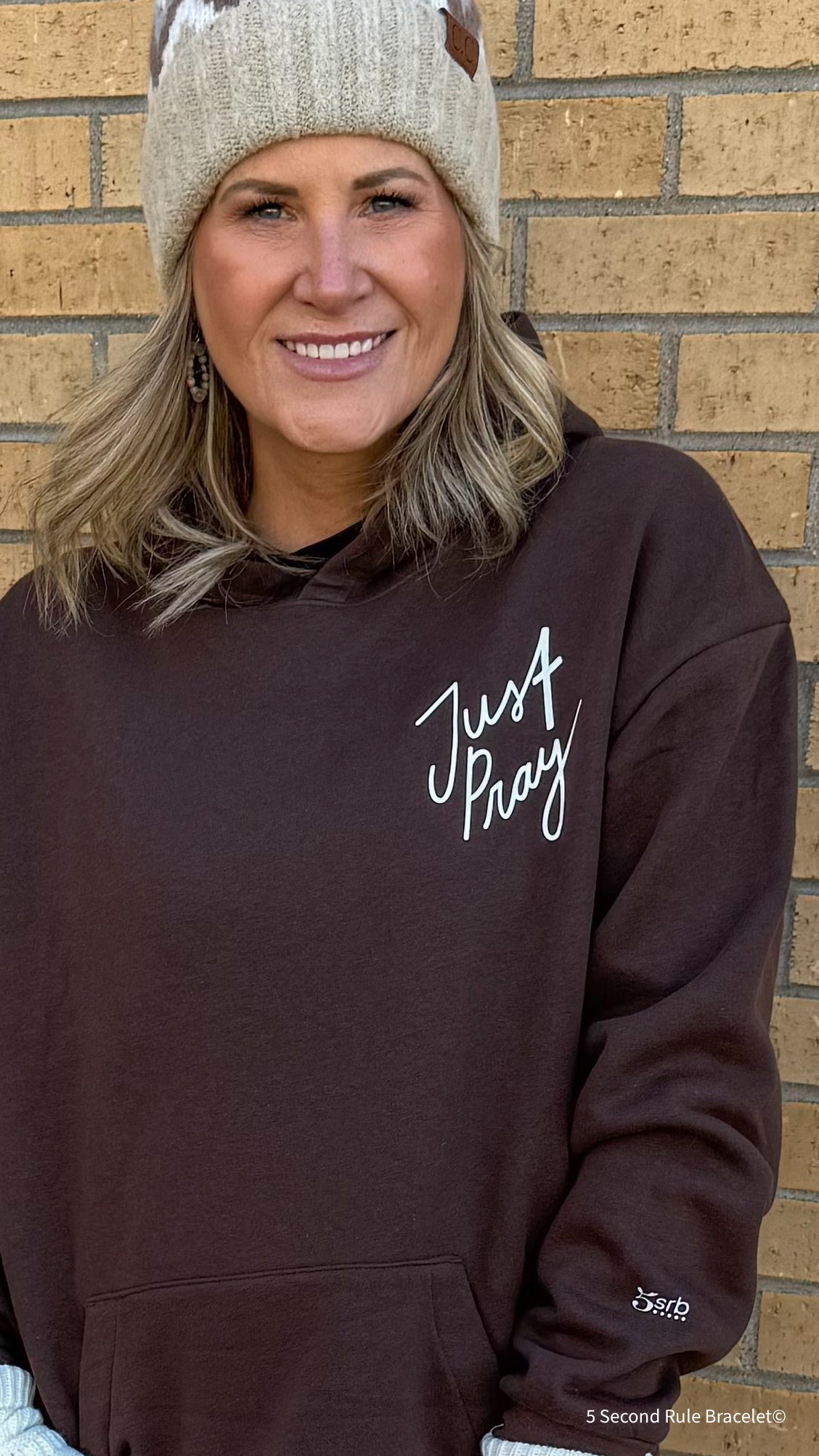 Woman wearing a brown hoodie with 'Just Pray' text against a brick wall.