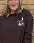 Woman wearing a brown hoodie with 'Just Pray' text against a brick wall.