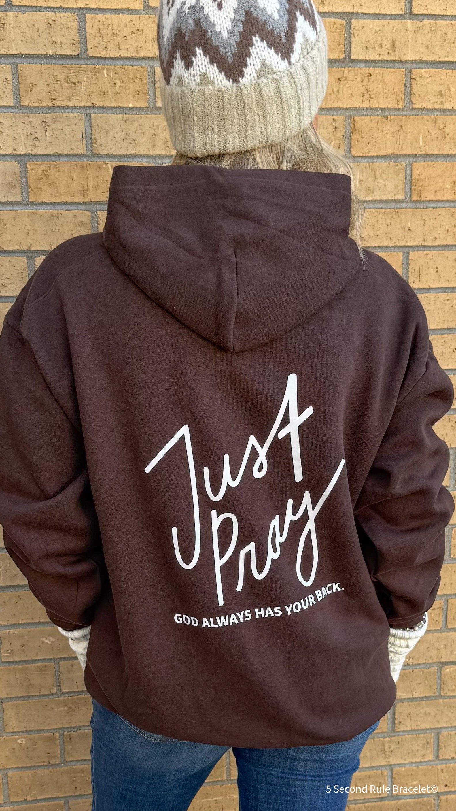 Person wearing a brown hoodie with 'Just Pray' text on the back against a brick wall.