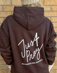 Person wearing a brown hoodie with 'Just Pray' text on the back against a brick wall.