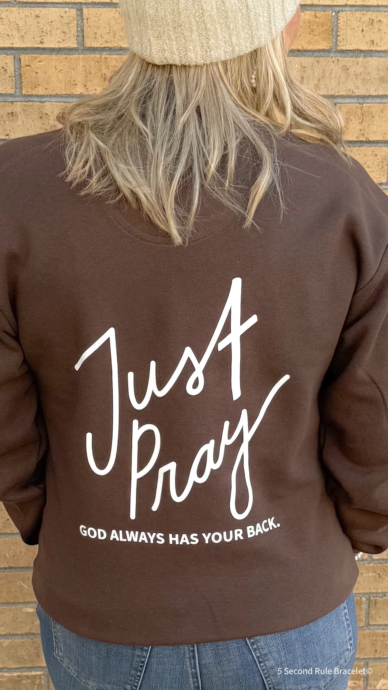 Brown sweatshirt with 'Just Pray' text on the back, worn by a person against a brick wall.