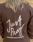 Brown sweatshirt with 'Just Pray' text on the back, worn by a person against a brick wall.
