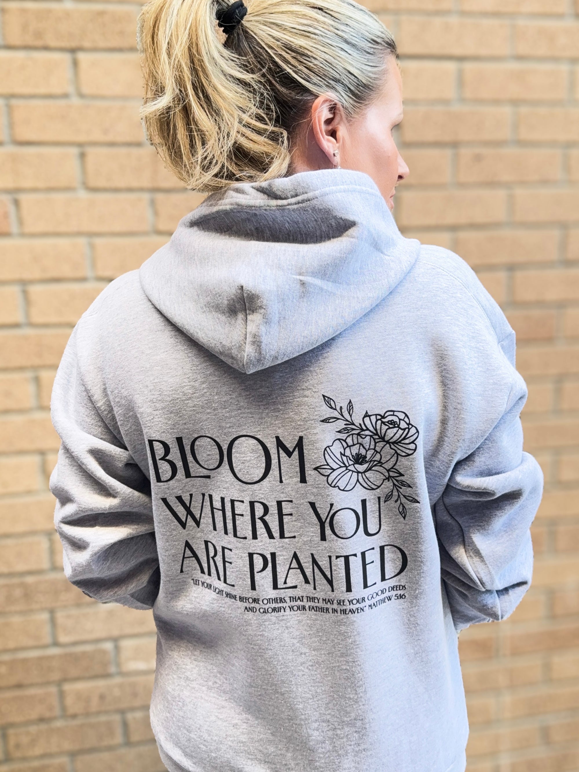Person wearing a gray hoodie with text and floral design on the back, standing against a brick wall.