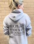 Person wearing a gray hoodie with text and floral design on the back, standing against a brick wall.