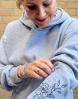 Person wearing a gray hoodie with a floral design and initials on a brick wall background