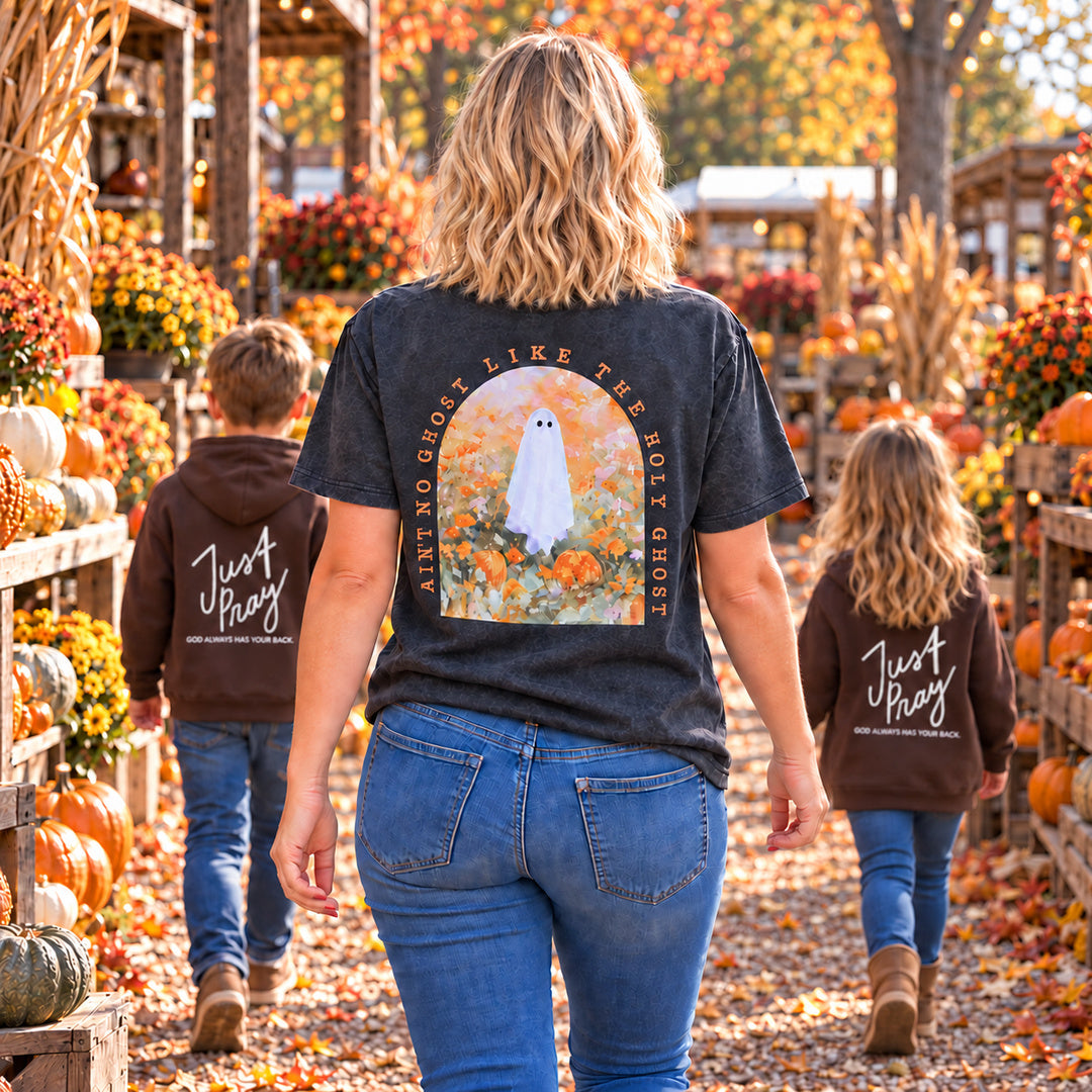 A woman and two children walk down a fall-themed pumpkin patch aisle, viewed from behind. The woman wears a dark charcoal graphic T-shirt with a ghost illustration and the words 'Ain't no ghost like the holy Ghosts,' while the children wear matching brown hoodies with 'Just Pray' written on the back.