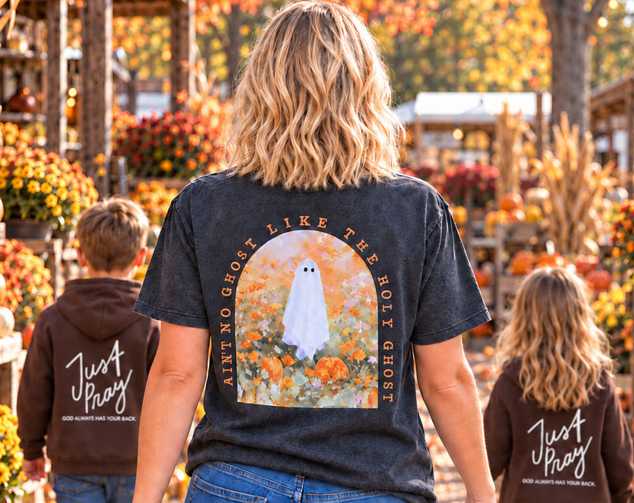 A woman and two children walk down a fall-themed pumpkin patch aisle, viewed from behind. The woman wears a dark charcoal graphic T-shirt with a ghost illustration and the words 'Ain't no ghost like the holy Ghosts,' while the children wear matching brown hoodies with 'Just Pray' written on the back.