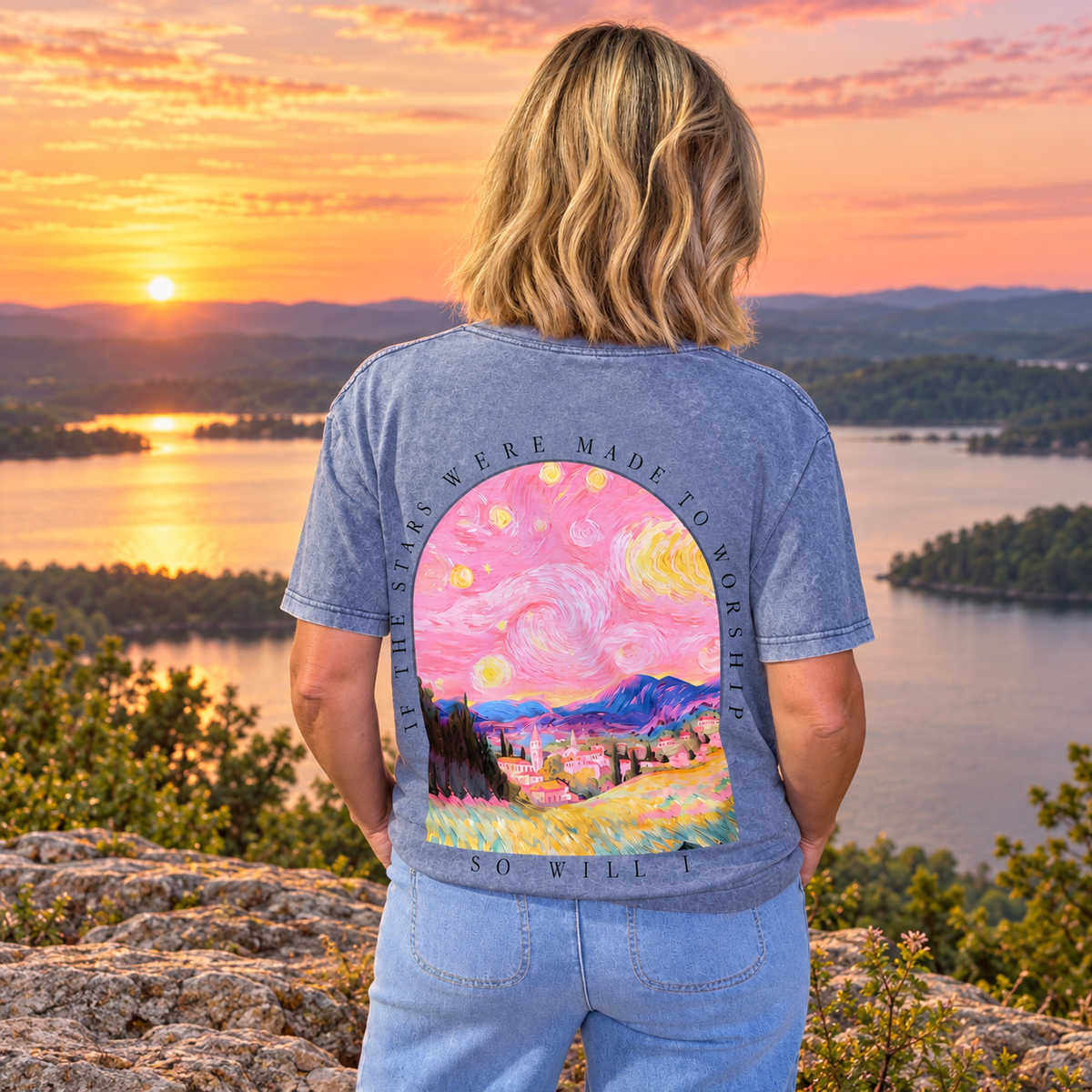 A woman stands on a rocky overlook at sunset with a lake and forested hills in the background, wearing a blue-gray graphic T-shirt from the back. The shirt features a large colorful print inspired by Van Gogh, with swirling pink and yellow sky, a village, and the text 'THE STARS WERE MADE TO WORSHIP SO WILL A'.