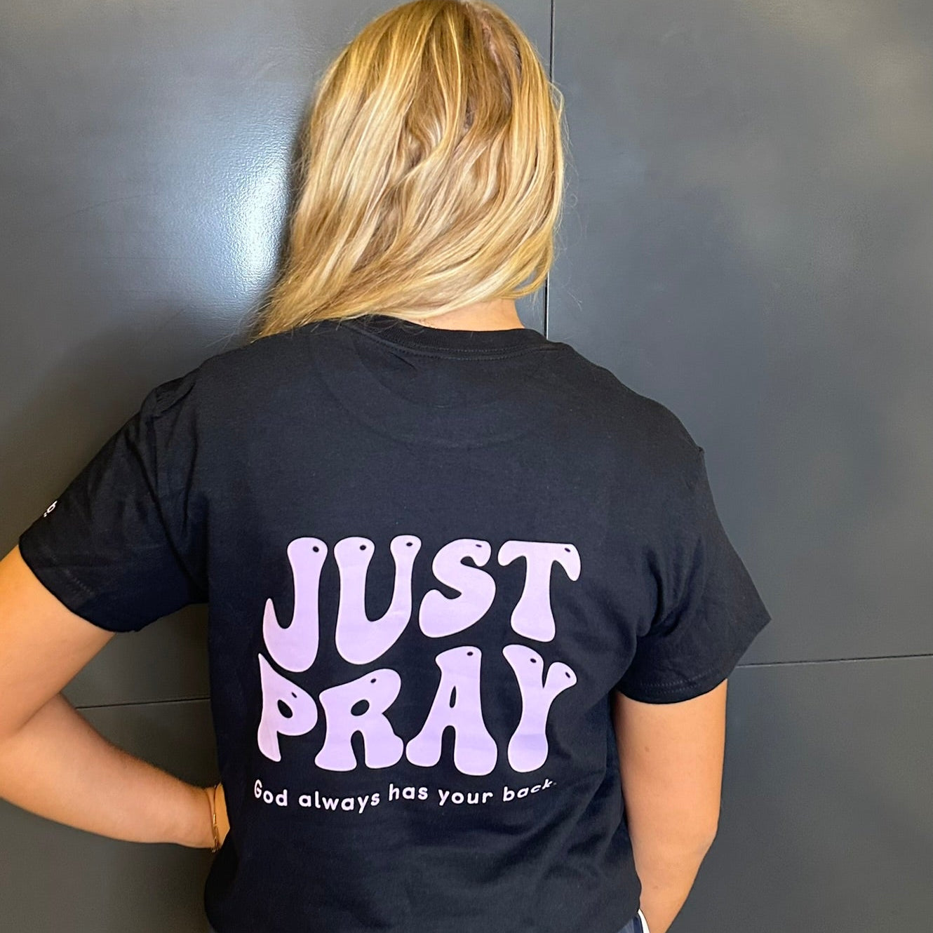 Person wearing a black t-shirt with 'JUST PRAY' text on the back against a gray wall.