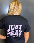 Person wearing a black t-shirt with 'JUST PRAY' text on the back against a gray wall.