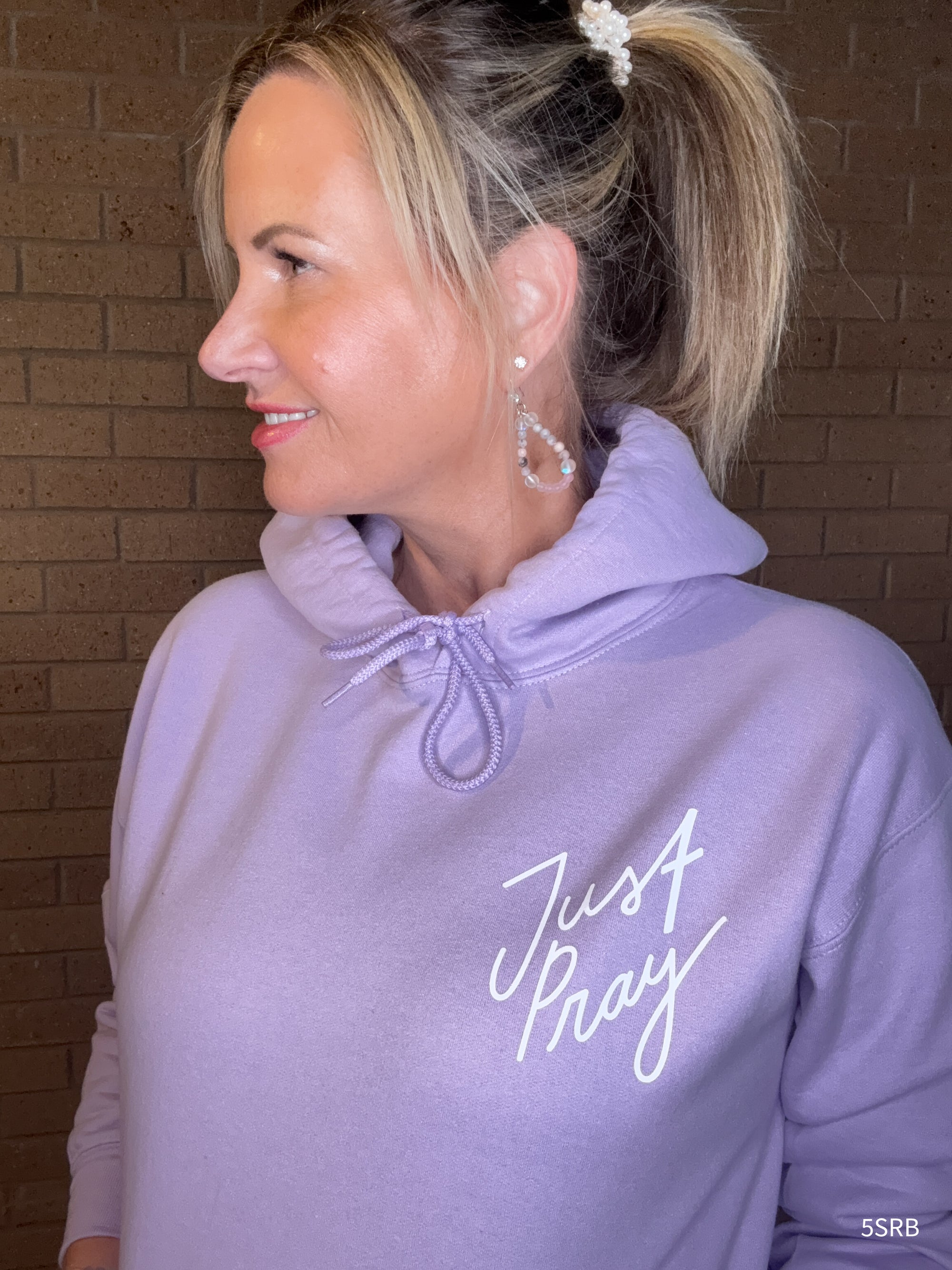 Woman wearing a lavender hoodie with 'Just Pray' text against a brick wall.