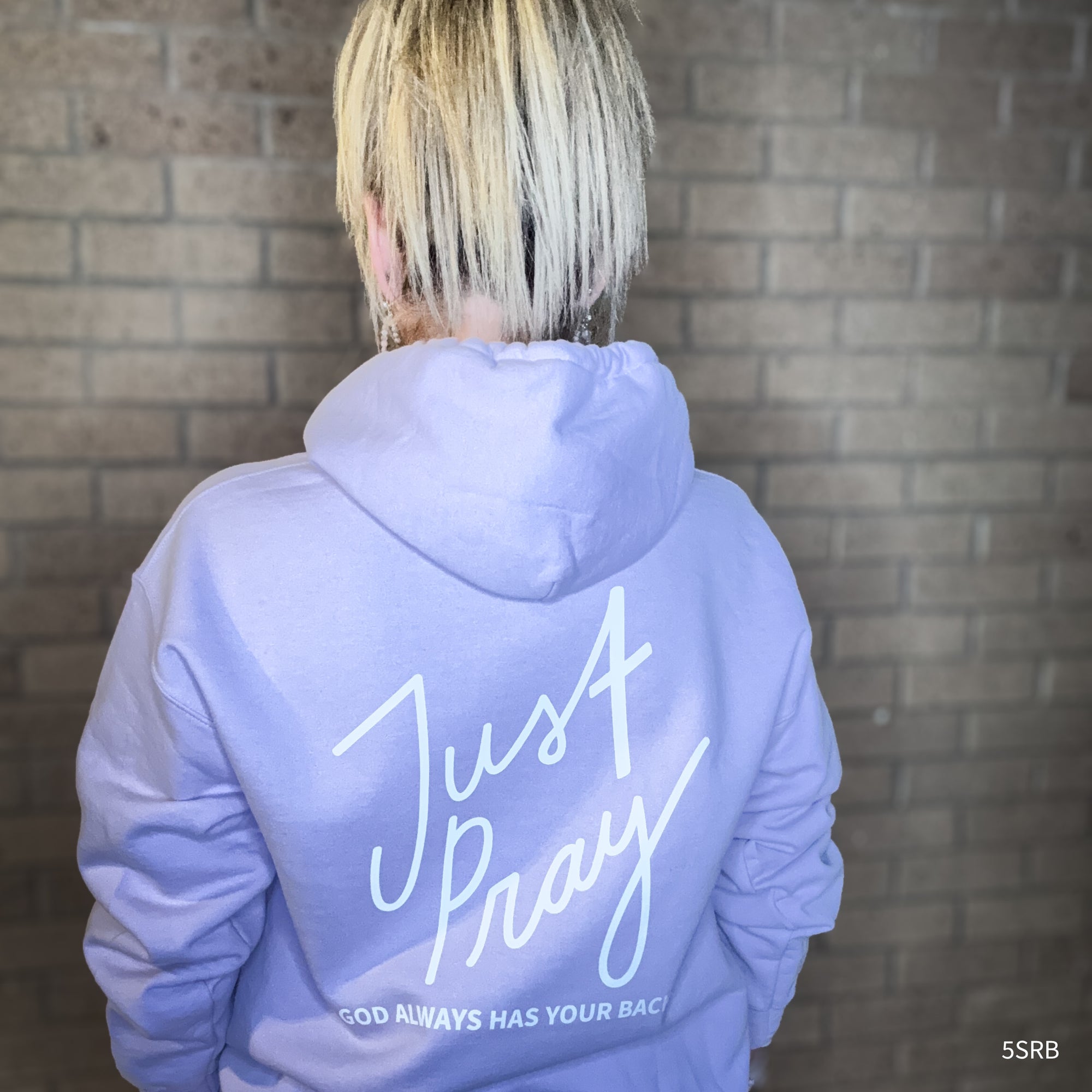 Person wearing a light blue hoodie with 'Just Pray' text on the back against a brick wall.