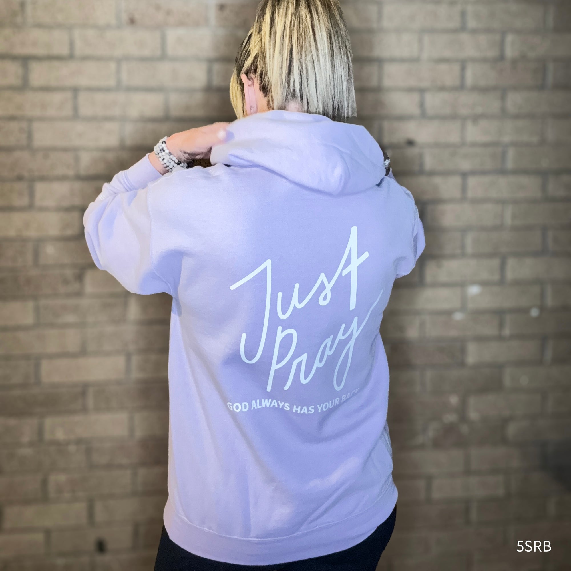 Person wearing a light purple hoodie with 'Just Pray' text on the back against a brick wall.