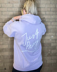 Person wearing a light purple hoodie with 'Just Pray' text on the back against a brick wall.