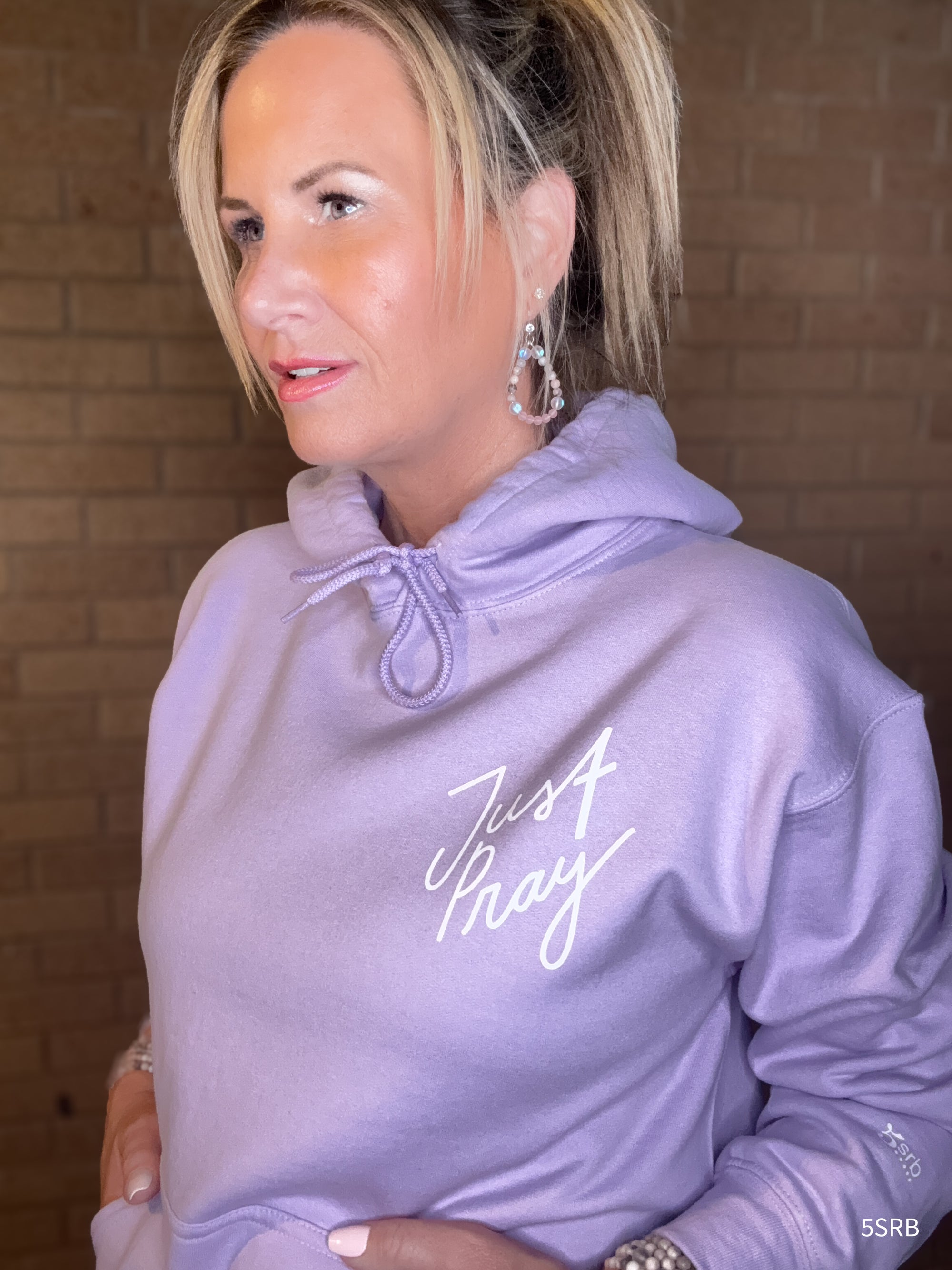Woman wearing a lavender hoodie with 'Just Pray' text against a brick wall.