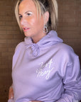 Woman wearing a lavender hoodie with 'Just Pray' text against a brick wall.