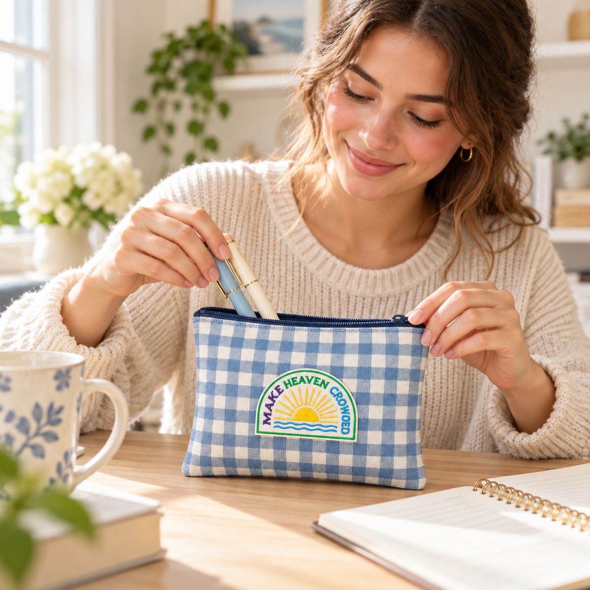 A woman in a cream knit sweater sits at a wooden desk, opening a blue and white gingham pencil case with a 'MAKE HEAVEN CLOUDS' sun logo. The desk is styled with a floral mug, books, and an open notebook.