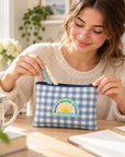A woman in a cream knit sweater sits at a wooden desk, opening a blue and white gingham pencil case with a 'MAKE HEAVEN CLOUDS' sun logo. The desk is styled with a floral mug, books, and an open notebook.