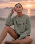 Woman wearing a 'Praise the Lord' sweatshirt sitting on sand with sunset in the background