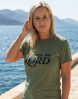 Woman wearing a green t-shirt with 'Praise the Lord' text by a body of water.