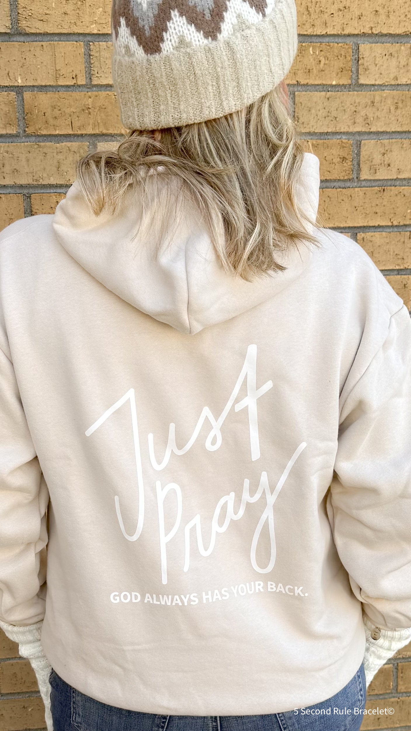 Person wearing a beige hoodie with 'Just Pray' text on the back against a brick wall.