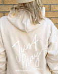 Person wearing a beige hoodie with 'Just Pray' text on the back against a brick wall.