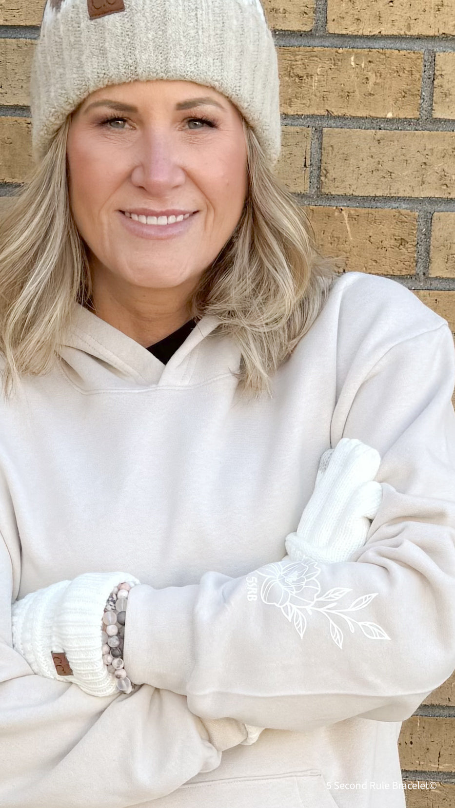 Person wearing a white beanie, white hoodie, and white gloves standing against a brick wall.
