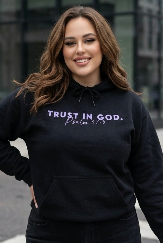 Woman wearing a black hoodie with 'Trust in God' text, standing outdoors.