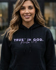 Woman wearing a black hoodie with 'Trust in God' text, standing outdoors.
