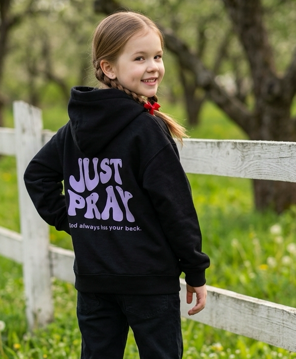 A young girl standing outdoors in a grassy field with a white fence and trees in the background, wearing a black hoodie with purple text reading 'JUST PRAY' on the back and black pants.