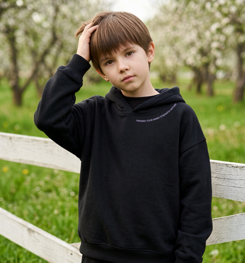 A young boy wearing a black pullover hoodie and matching black sweatpants stands in a grassy field with a white wooden fence and blooming trees in the background.