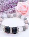 A Lava Stone and Rhinestone stretch bracelet made with 2 rough black lava stones, translucent aura beads, pink zebra jasper gemstones, and rhinestone accents for added sparkle sits on a white table. 