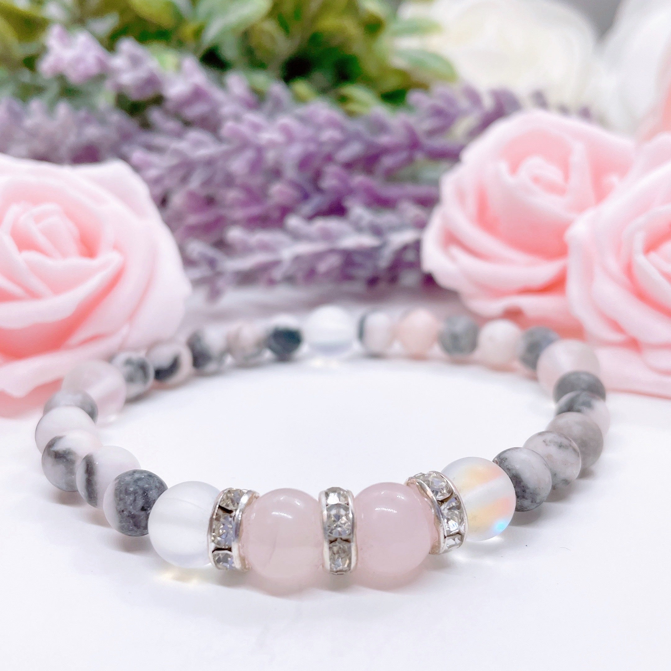 A Rose Quartz Rhinestone Companion Gemstone stretch bracelet made with 2  pink rose quartz gemstones, translucent aura beads, pink jasper gemstones, and rhinestone accents for added sparkle sits on a white table. 