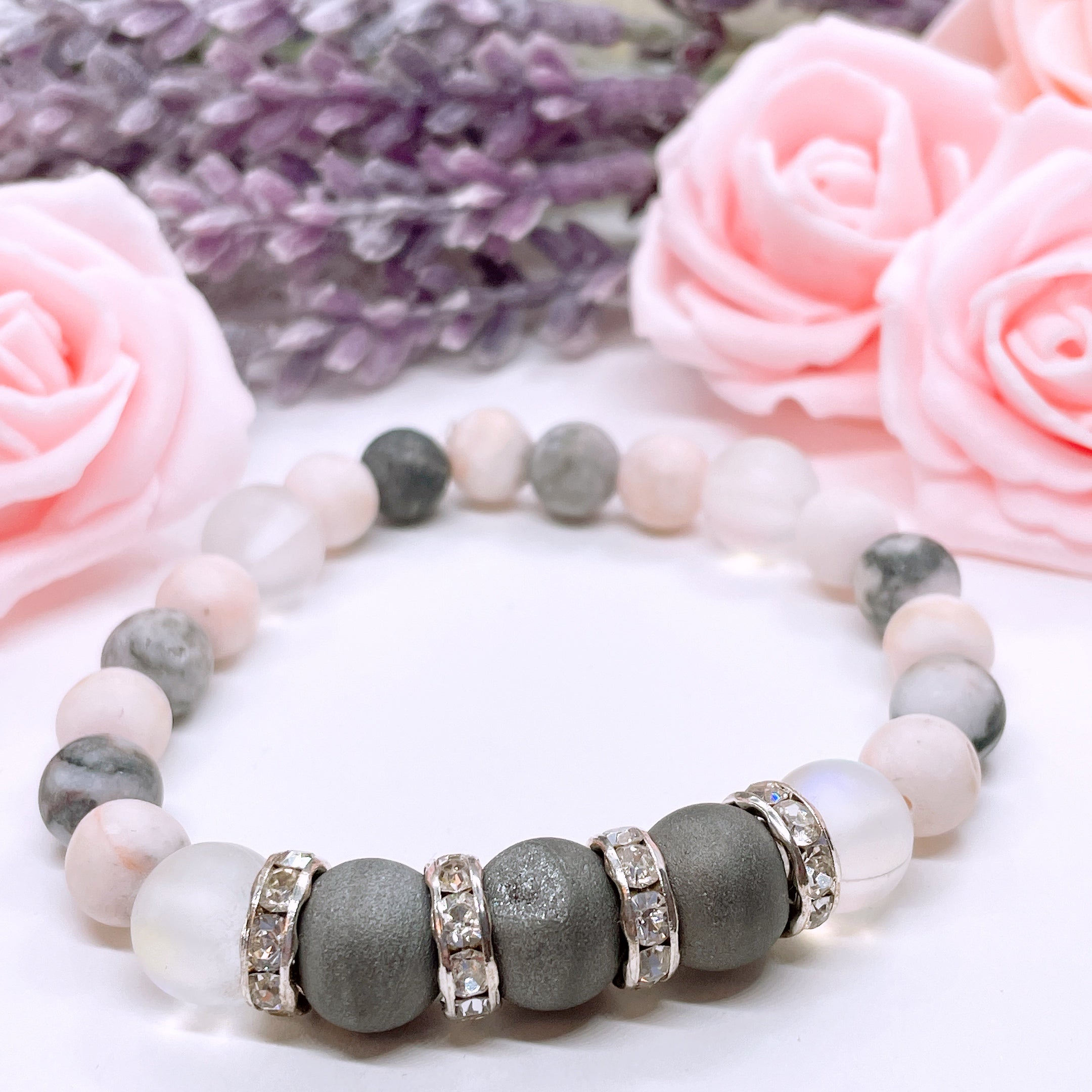 A Druzy Rhinestone stretch bracelet made with 3 dark rough druzy gemstones, translucent aura beads, pink zebra jasper gemstones, and rhinestone accents for added sparkle sits on a white table. 