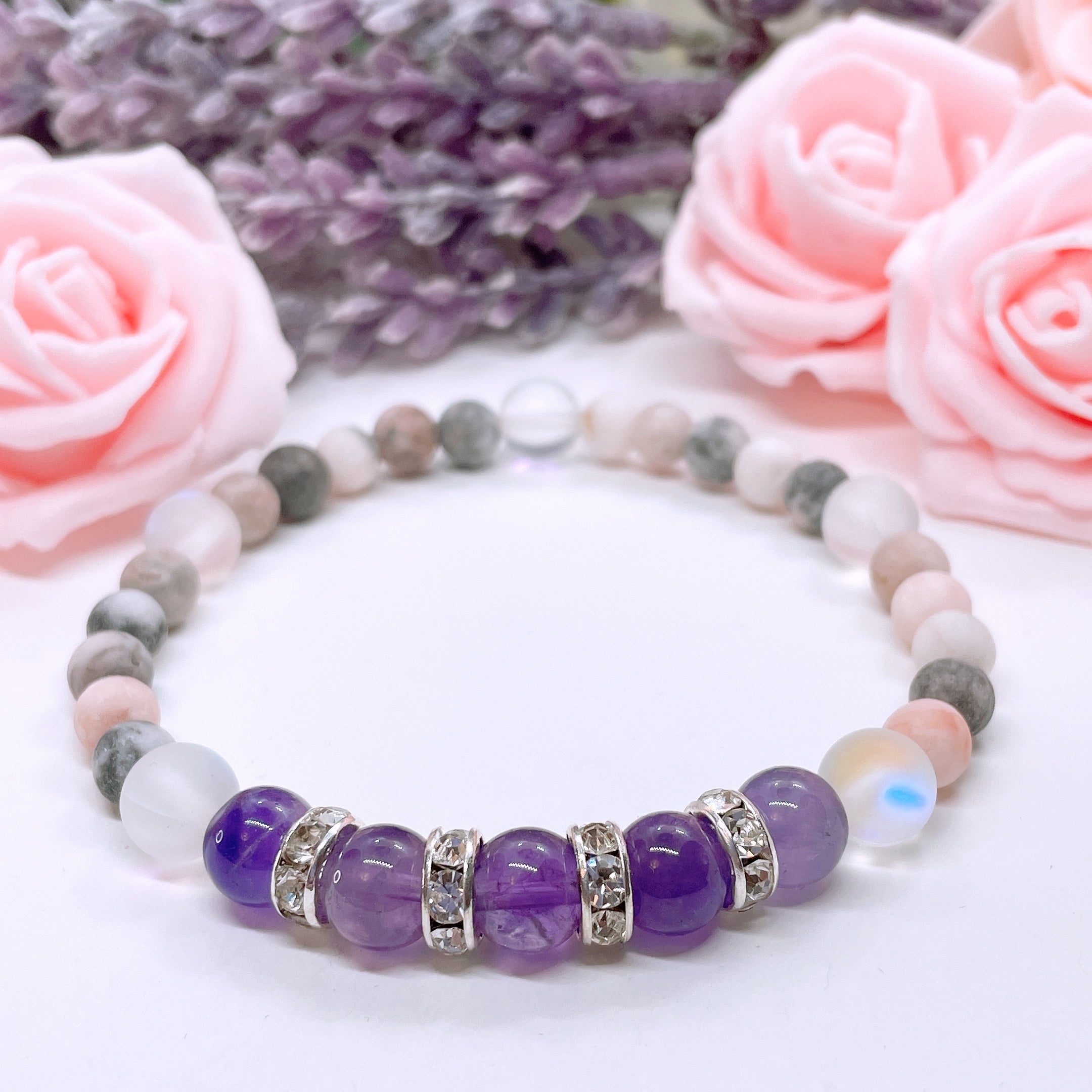 The Amethyst Rhinestone Companion Gemstone stretch bracelet made with 5 dark purple amethyst gemstones, translucent aura beads, and rhinestone accents for added sparkle sits on a white table. 