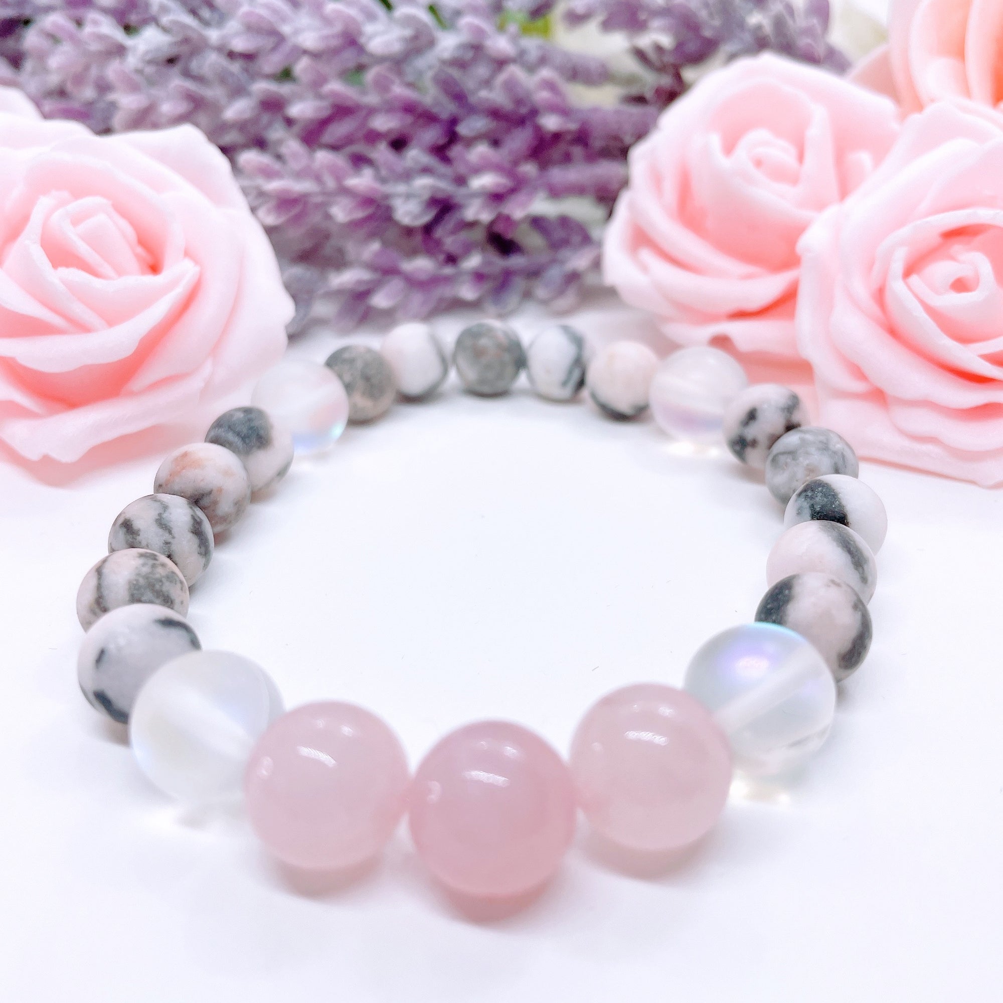 A Rose Quartz Gemstone stretch bracelet made with 2 pink rose quartz gemstones, translucent aura beads, and pink jasper gemstones sit on a white table. 