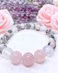 A Rose Quartz Gemstone stretch bracelet made with 2 pink rose quartz gemstones, translucent aura beads, and pink jasper gemstones sit on a white table. 