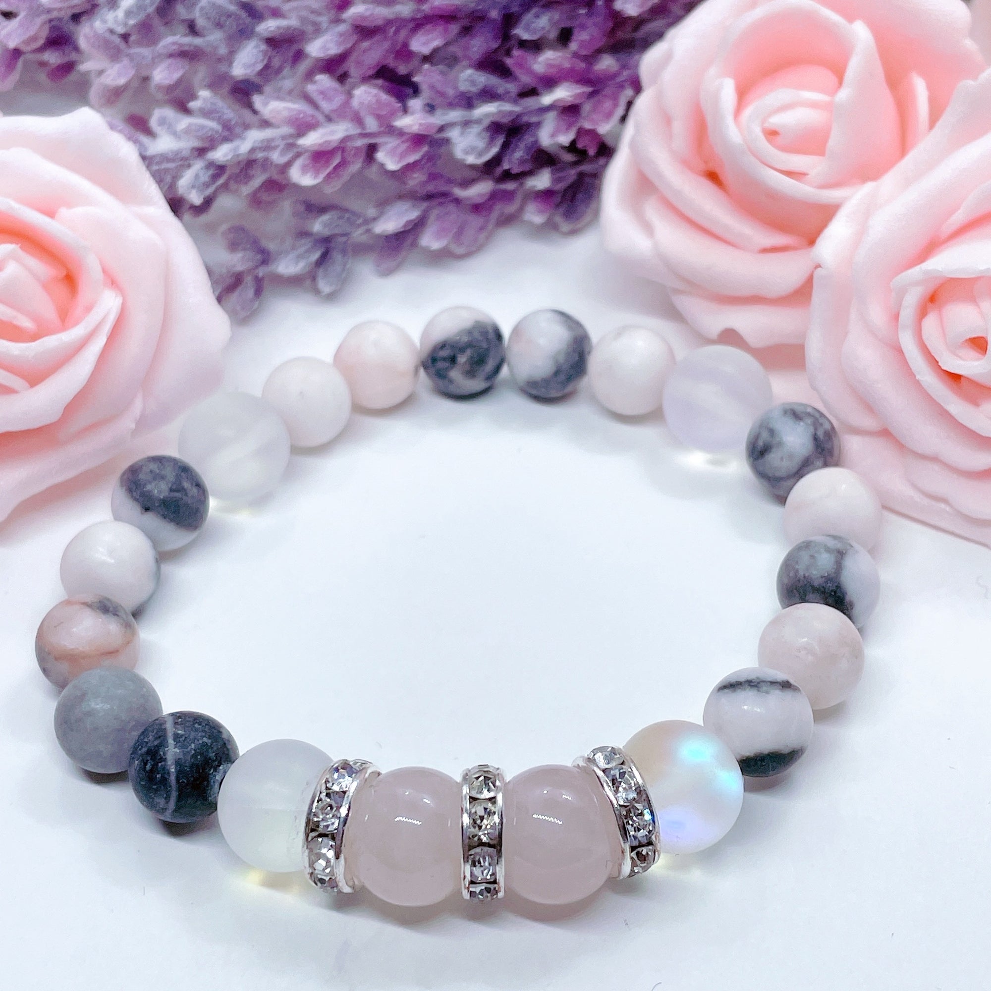 A Rose Quartz Rhinestone and Gemstone stretch bracelet made with 2  pink rose quartz gemstones, translucent aura beads, pink jasper gemstones, and rhinestone accents for added sparkle sits on a white table. 