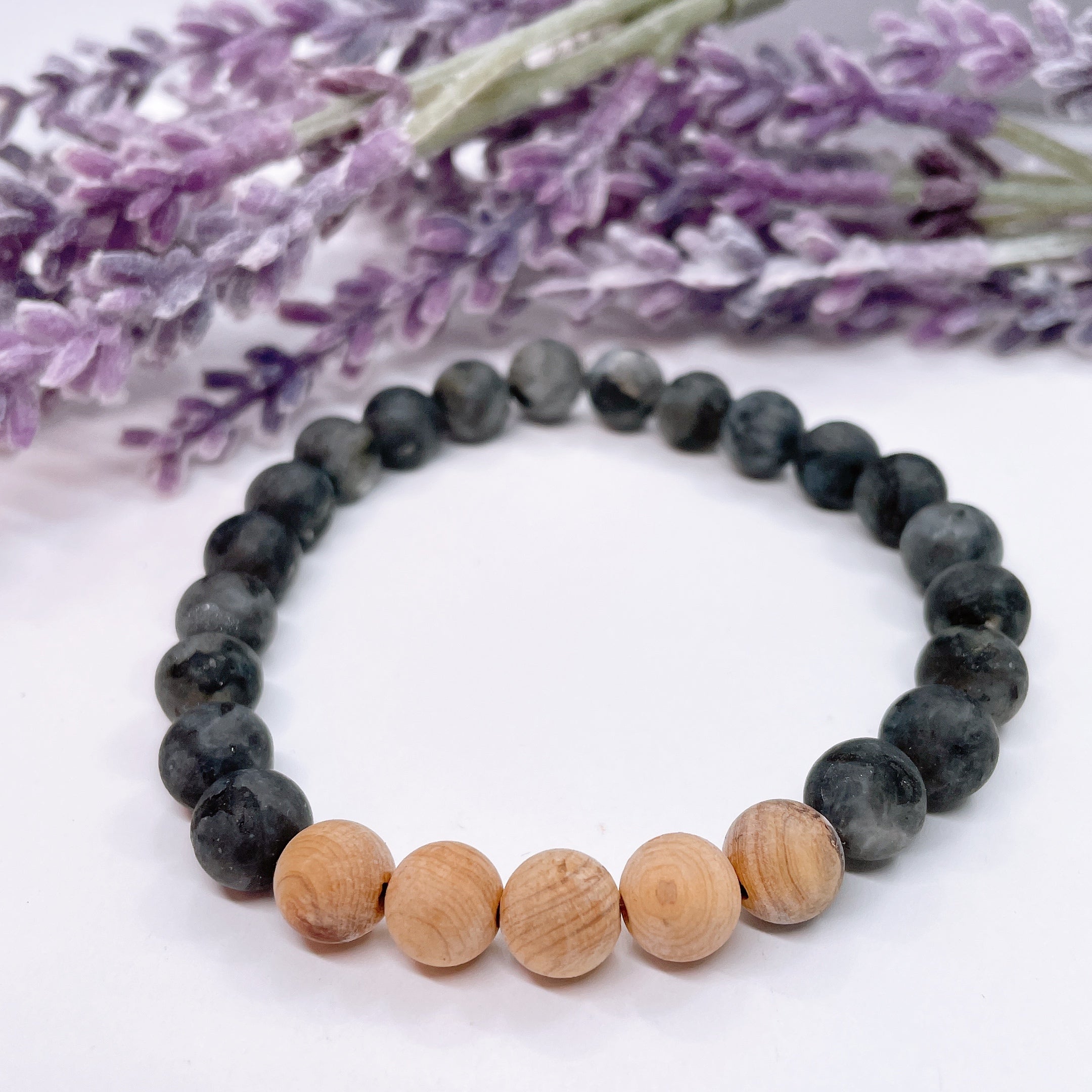 A black beaded bracelet with 20 black labradorite beads with 5 cedar wood brown beads on a white table with a purple flower on the table.