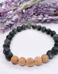 A black beaded bracelet with 20 black labradorite beads with 5 cedar wood brown beads on a white table with a purple flower on the table.