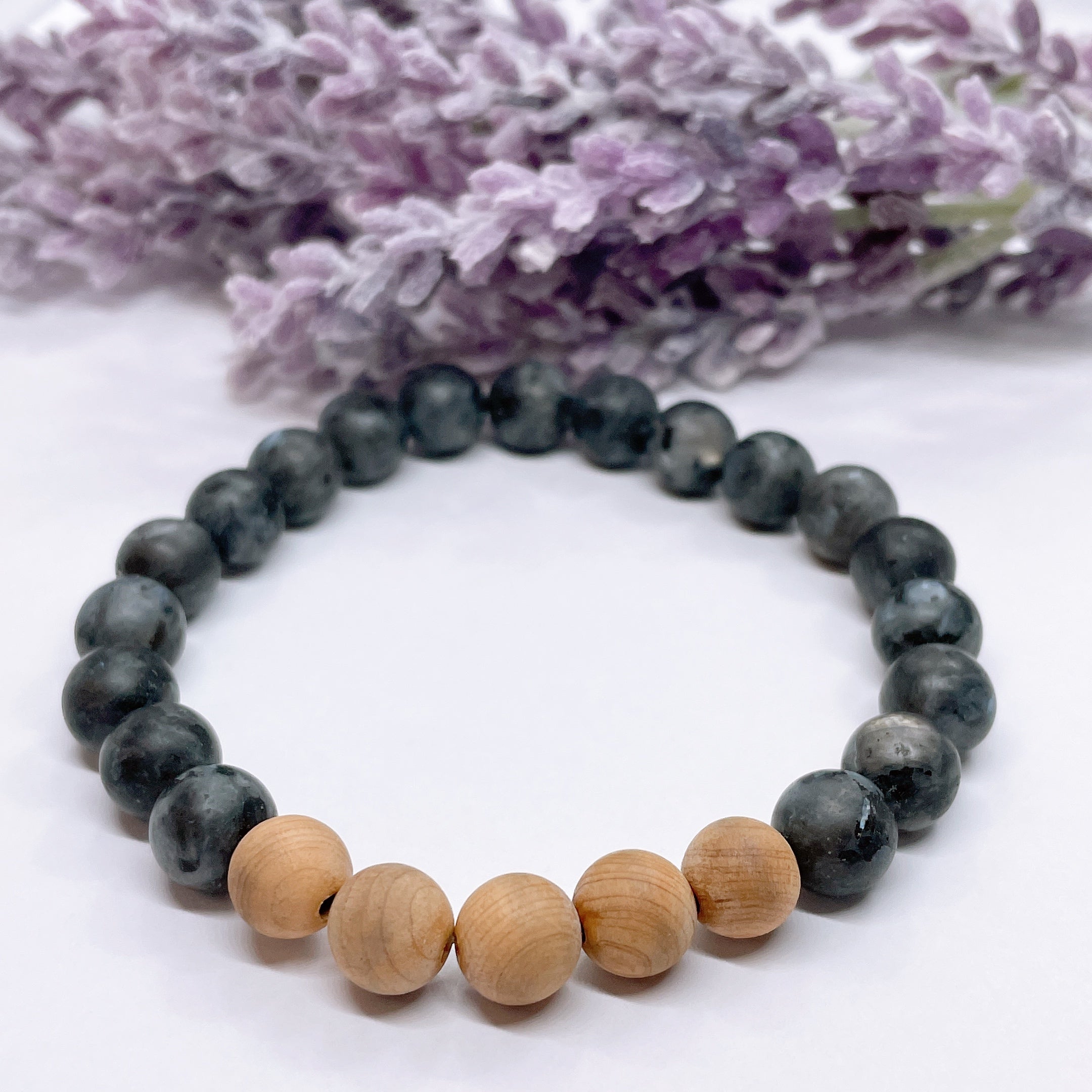 A black beaded bracelet with 19 black labradorite beads with 5 cedar wood brown beads on a white table with a purple flower on the table.