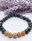 A black beaded bracelet with 19 black labradorite beads with 5 cedar wood brown beads on a white table with a purple flower on the table.