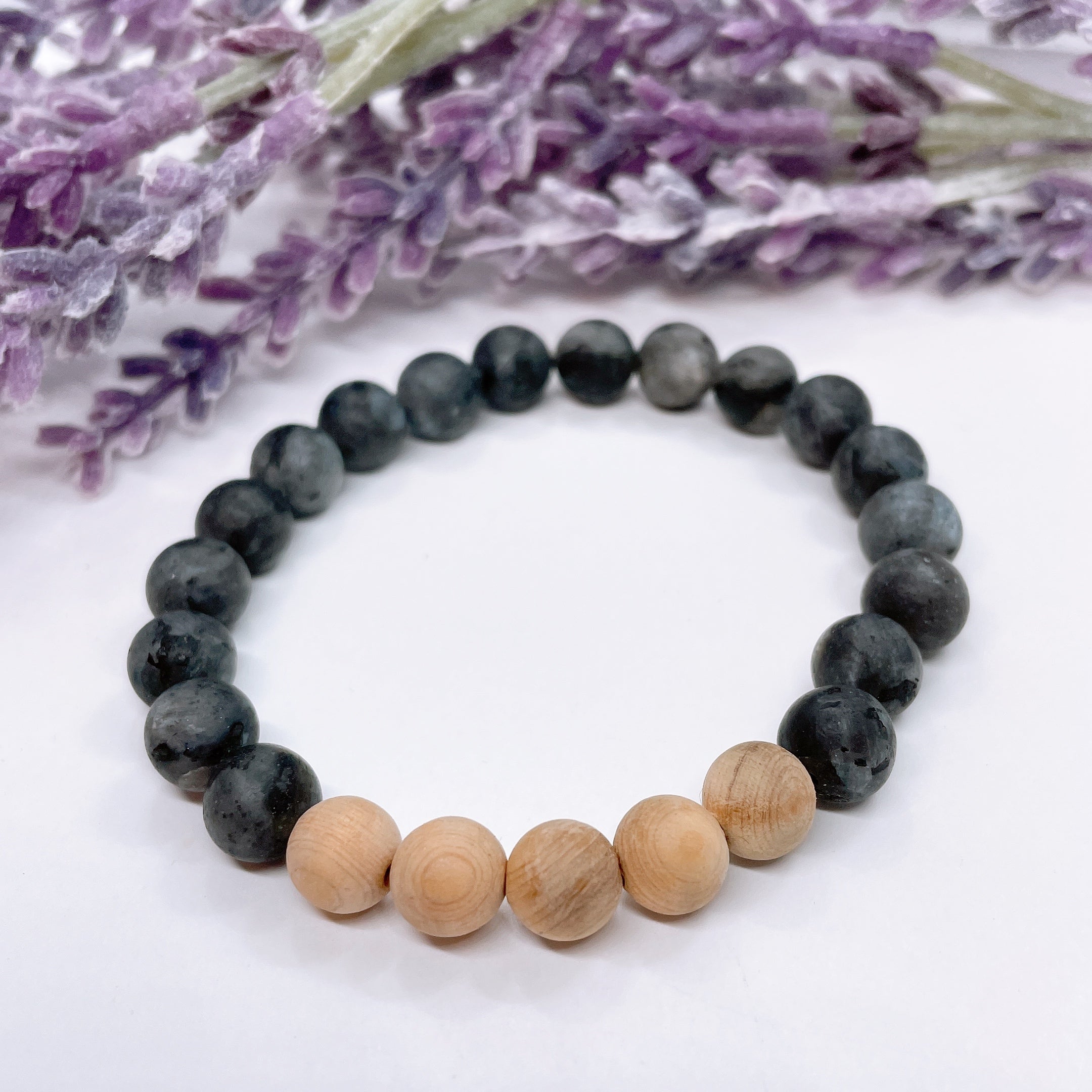 A black beaded bracelet with 18 black labradorite beads with 5 cedar wood brown beads on a white table with a purple flower on the table.