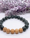 A black beaded bracelet with 16 black labradorite beads with 5 cedar wood brown beads on a white table with a purple flower on the table.
