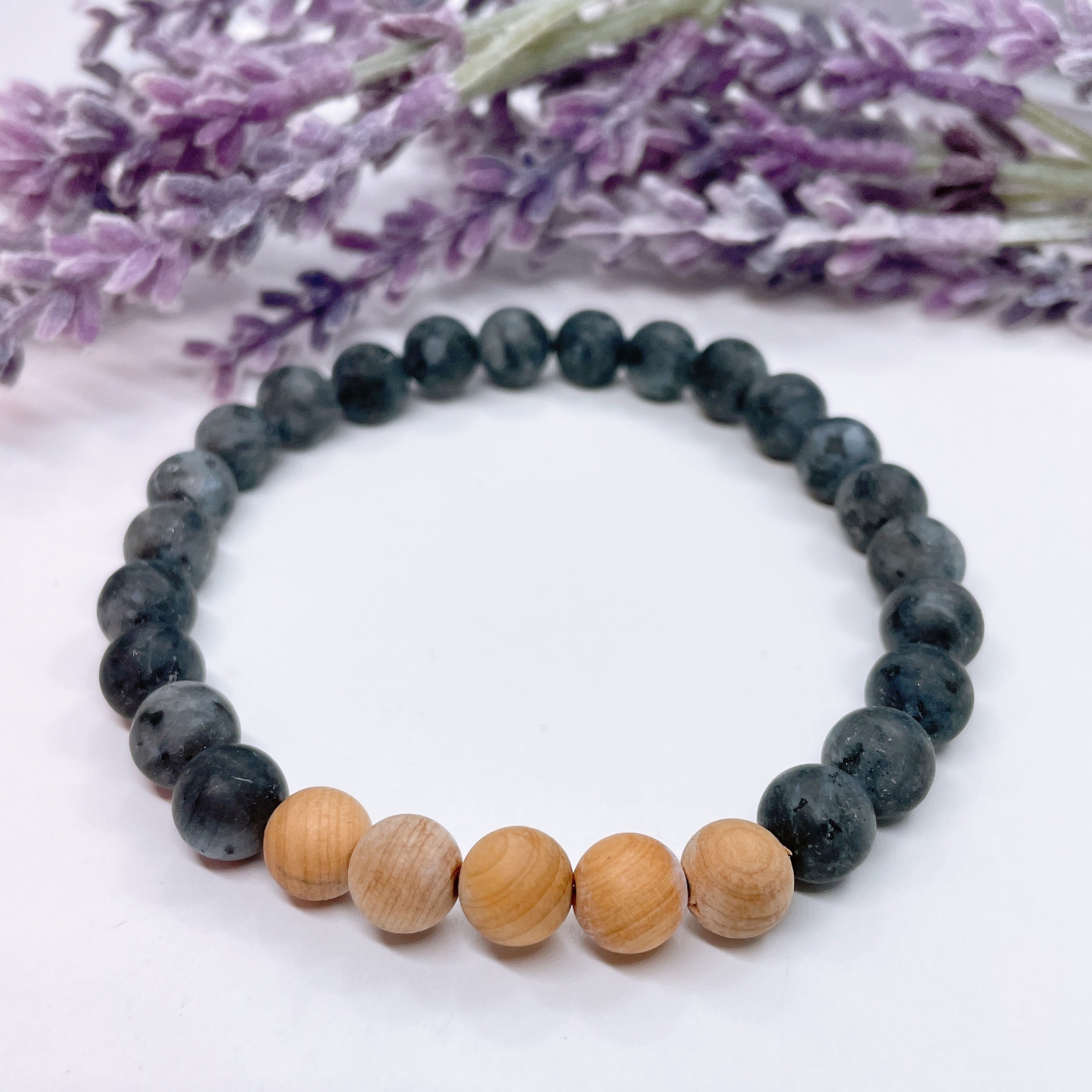 A black beaded bracelet with 22 black labradorite beads with 5 cedar wood brown beads on a white table with a purple flower on the table.