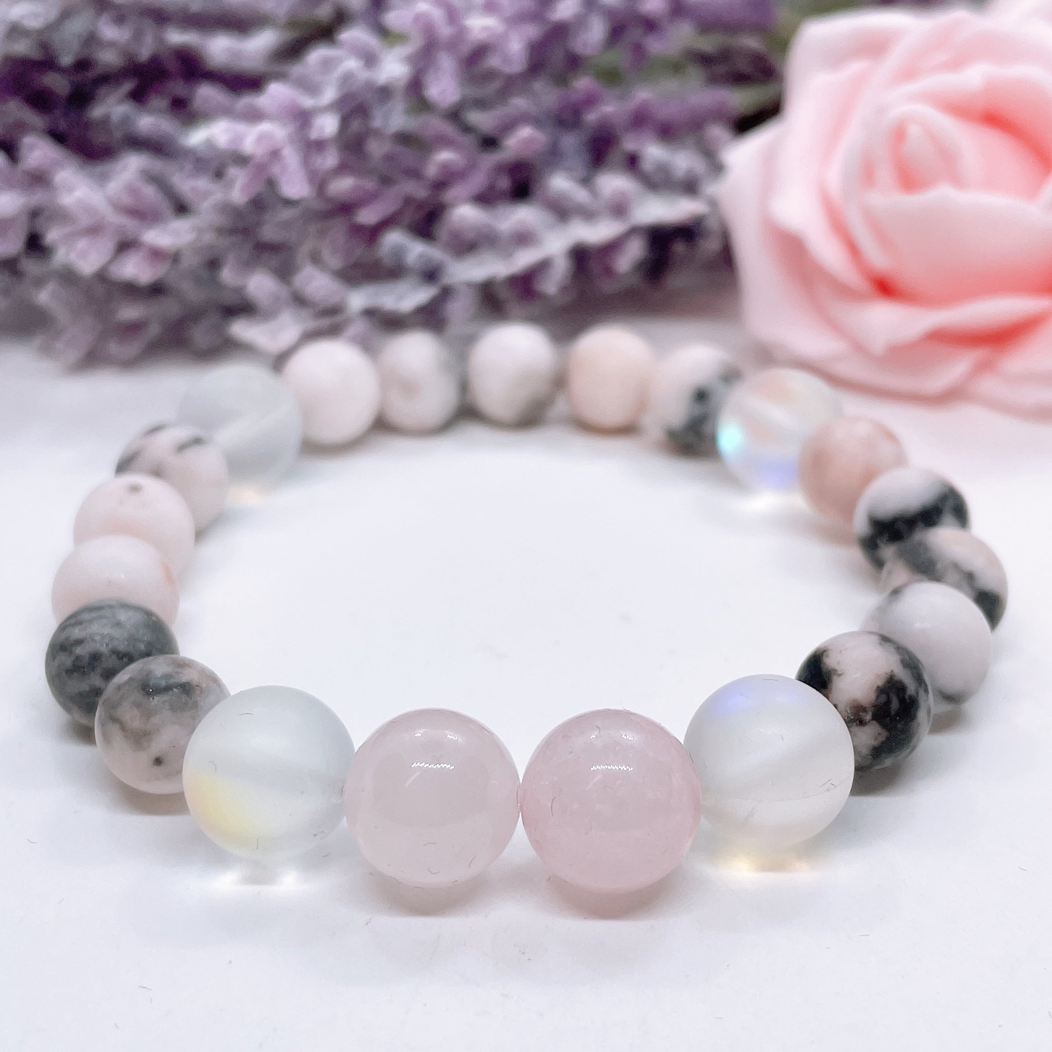 A Rose Quartz Gemstone stretch bracelet made with 2 pink rose quartz gemstones, translucent aura beads, and pink jasper gemstones sit on a white table. 